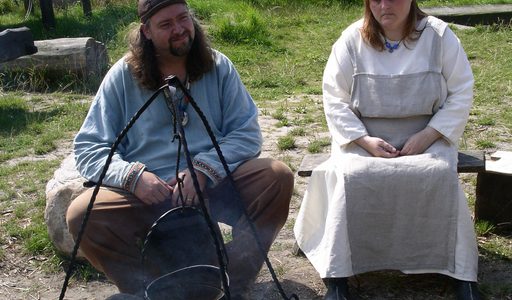 "Two Viking re-enactors in traditional summer day-wear garb tend a cook fire; Foteviken, Sweden."
