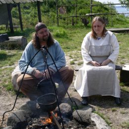 "Two Viking re-enactors in traditional summer day-wear garb tend a cook fire; Foteviken, Sweden."