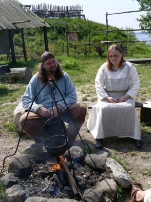 "Two Viking re-enactors in traditional summer day-wear garb tend a cook fire; Foteviken, Sweden."